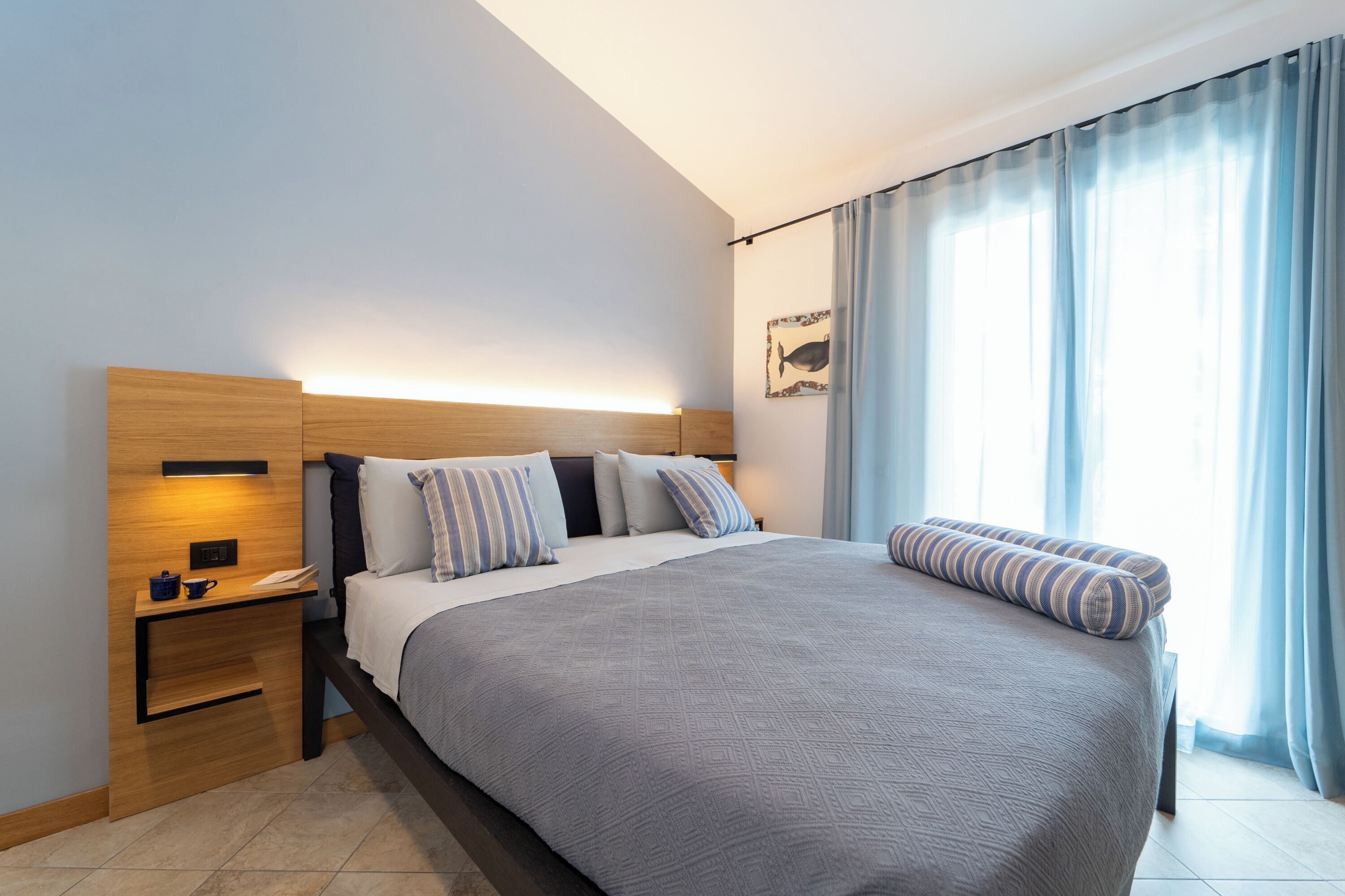 A modern hotel room with a large bed, a bedside table with a lamp, a small shelf, and a window with curtains.