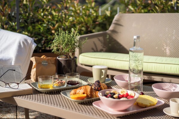 A table set with breakfast items including croissants, fruits, and beverages, with a green sofa and plants in the background. - Elbitat Homes (Rio)