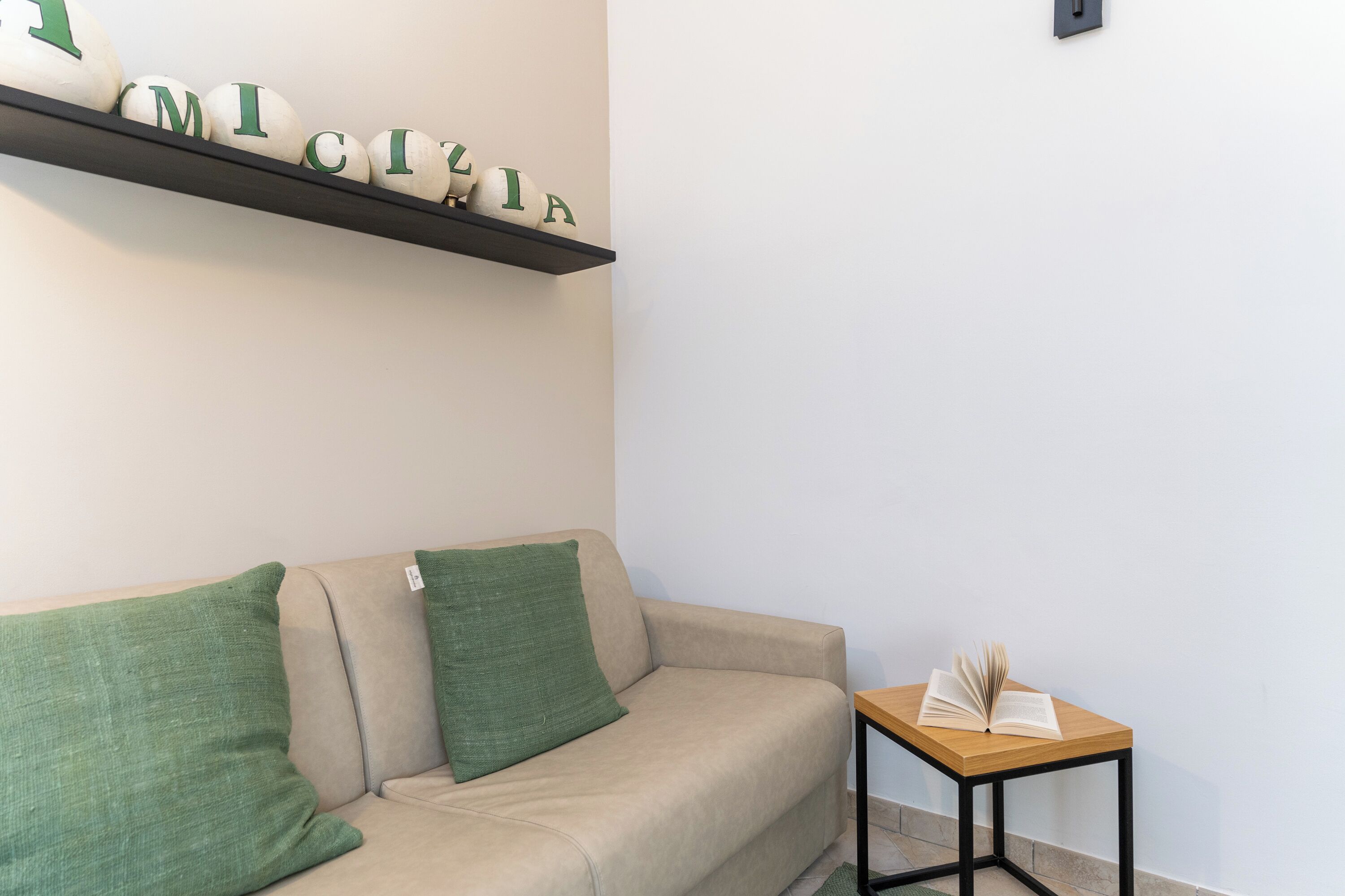 Ein beiges Sofa mit grünen Kissen, ein Holztischchen mit einem offenen Buch und ein Regal mit weißen Lettern, die "MICELIA" schreiben.