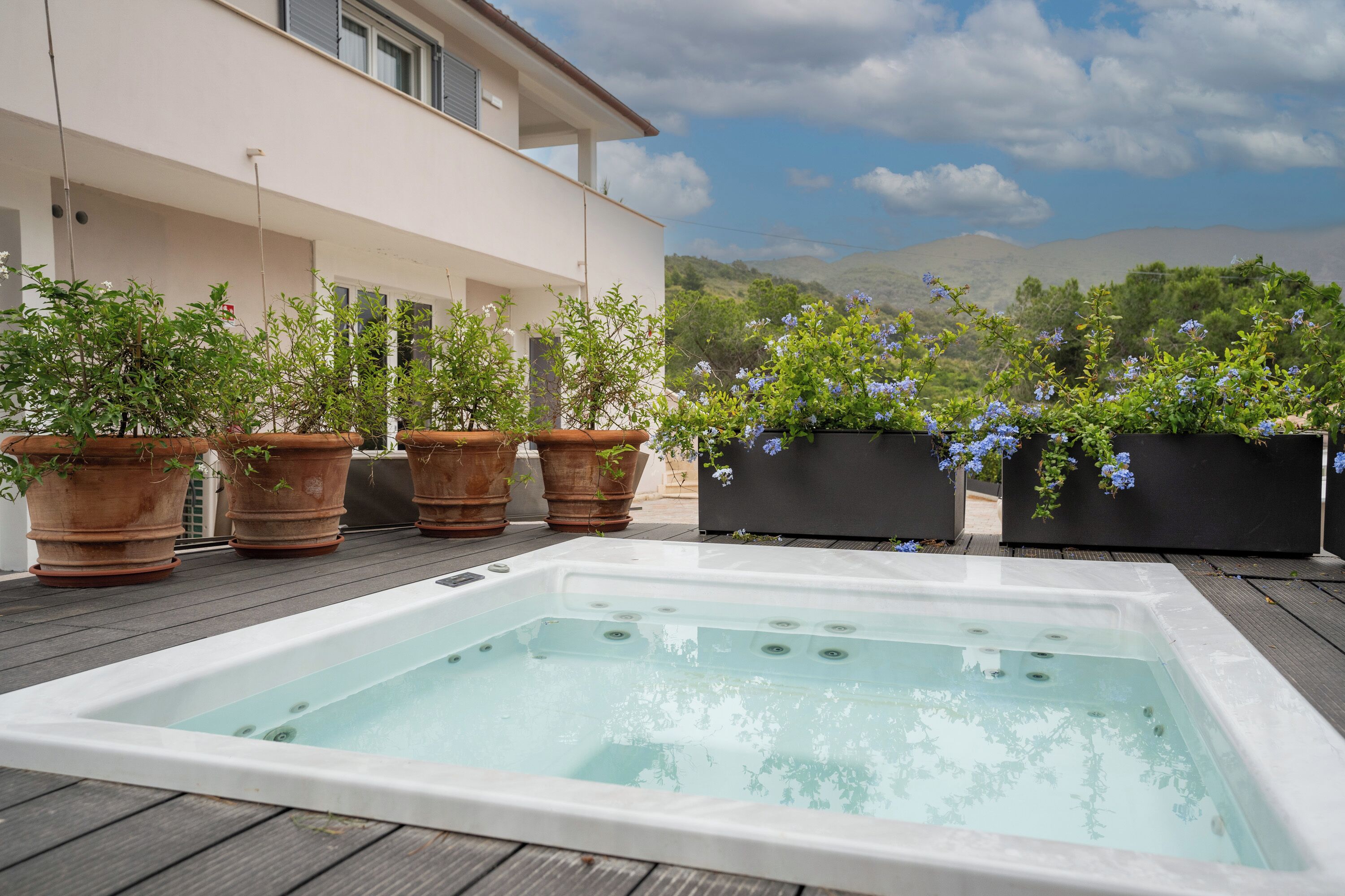 A hot tub on a wooden deck with potted plants and a building in the background.