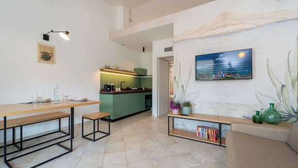 A modern kitchen with a green island, a dining area with a wooden table and benches, and a living area with a sofa and a wall-mounted TV. - Elbitat Homes (Rio)
