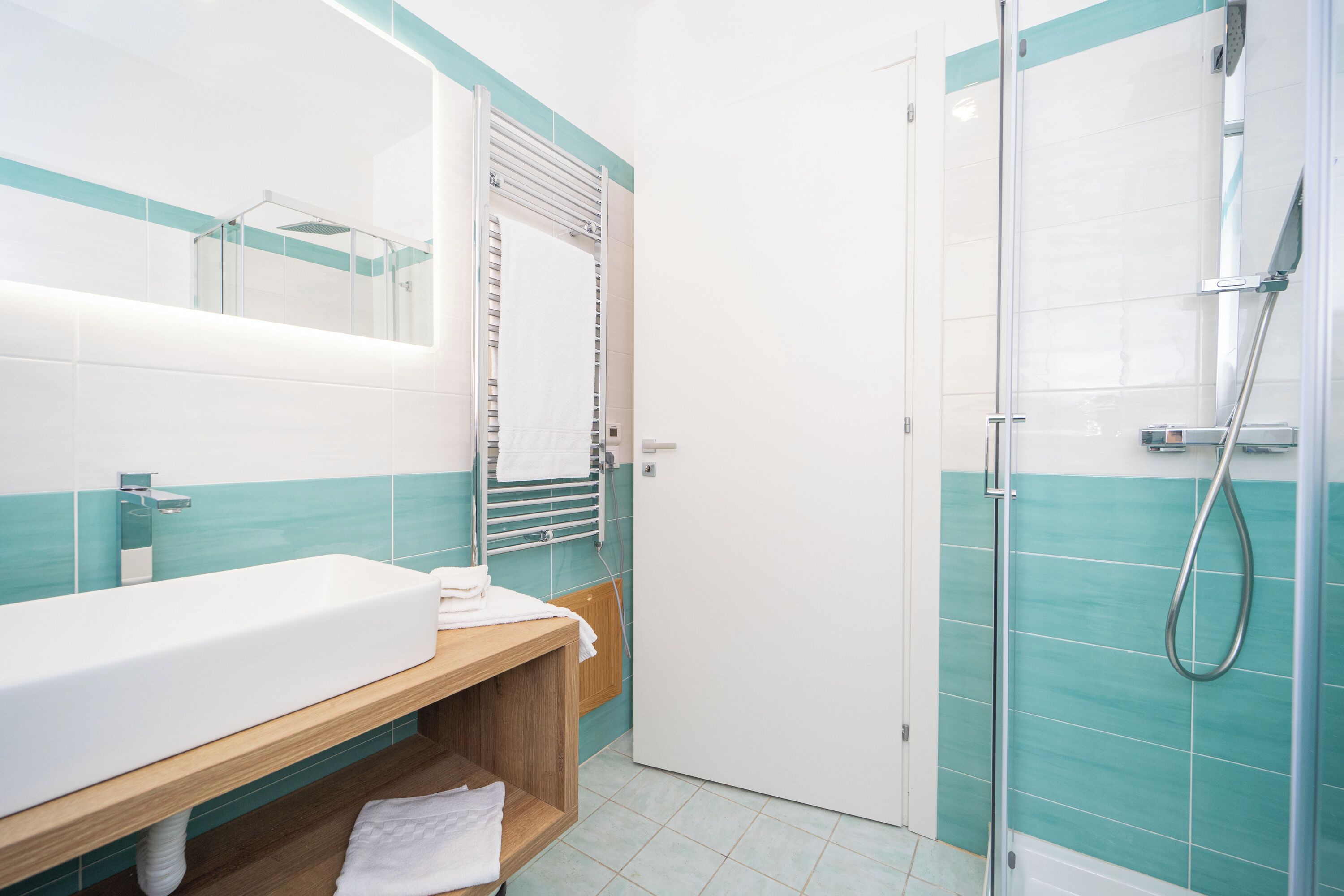 A modern bathroom with a large white sink, a wooden vanity, a glass shower enclosure, and teal and white tiled walls.