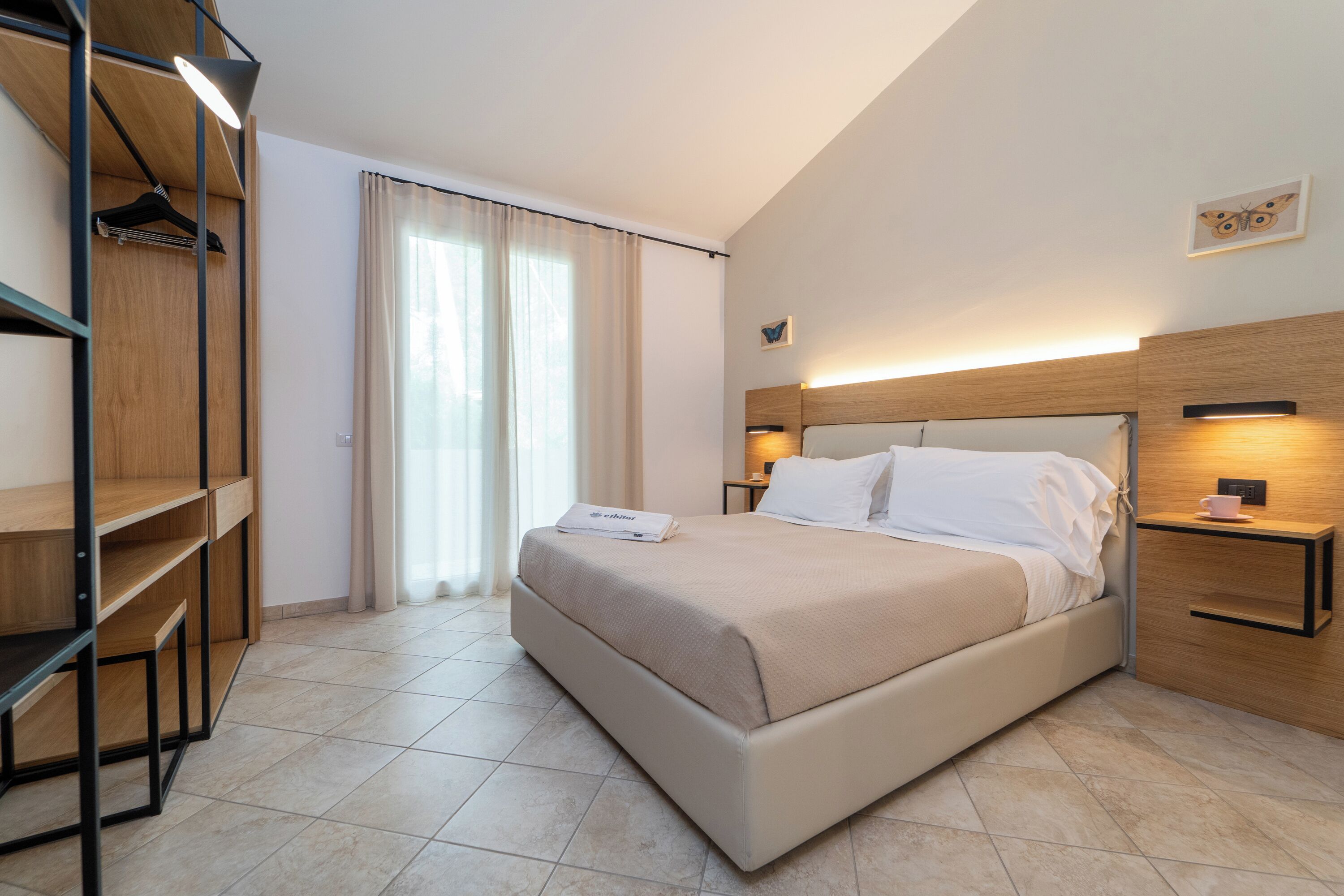 Ein modernes Hotelzimmer mit einem großen Bett, einem Holzschrank, einem Nachttisch mit Lampe und einem Fenster mit Vorhängen.