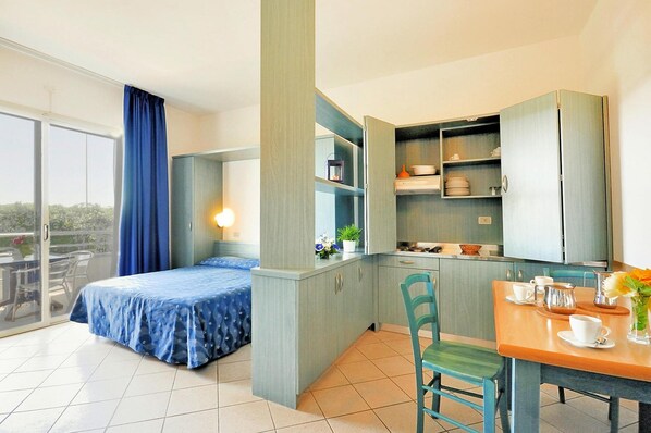 A compact room with a bed, a dining table with chairs, a kitchenette, and a sliding glass door leading to an outdoor area.