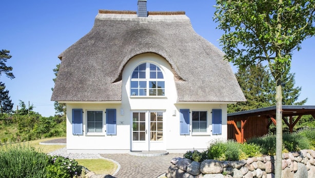 Ein Haus mit einem Strohdach, weißen Wänden und blauen Fensterläden. Es besitzt ein zentrales Rundbogenfenster und eine Mauer aus Stein davor.