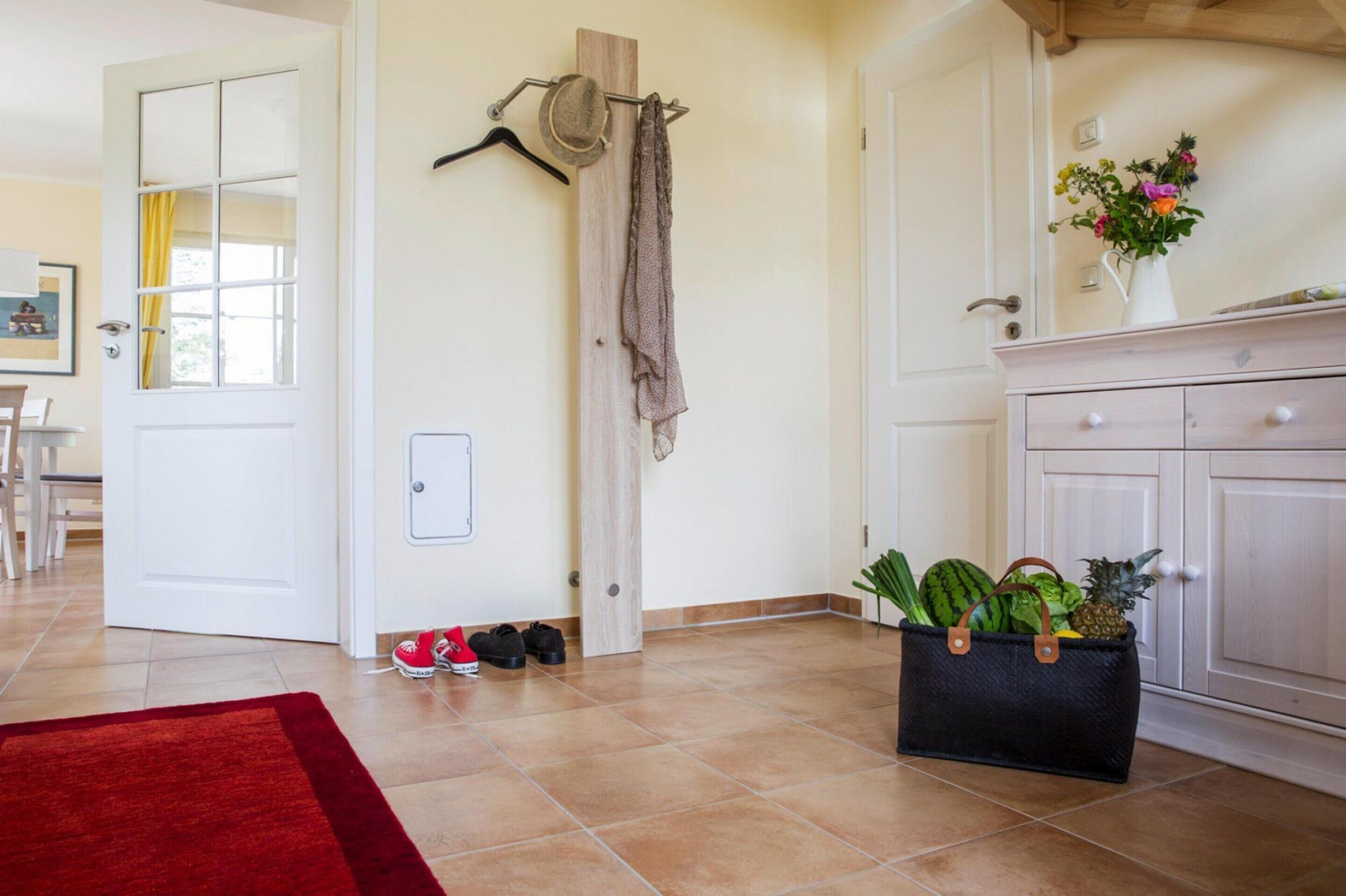 Une pièce avec un tapis rouge, une porte en bois, un porte-manteau avec des chapeaux, une armoire ornée d’un vase de fleurs et un panier rempli de fruits.