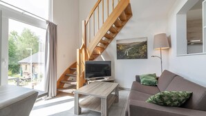 A modern living room with a wooden staircase, a flat-screen TV, a sofa with green cushions, and a large window offering a view of the outdoors. - Shared Pool and Sauna, Mistelgau-obernsees (Mistelgau)
