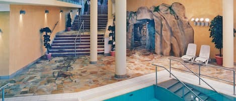 An indoor swimming pool with stone flooring, a staircase, and a rock wall mural.