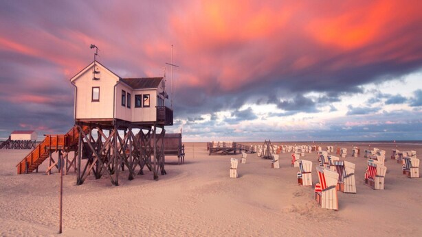 Una casa sulla spiaggia con una veranda in legno e una torretta per i bagnini, circondata da sdraio e ombrelloni.