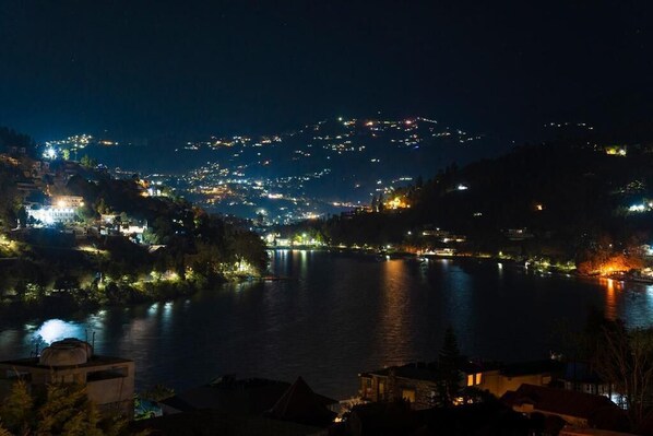 A nighttime view of a riverside town with illuminated buildings and a calm river. - HR Upvan Inn (Nainital)