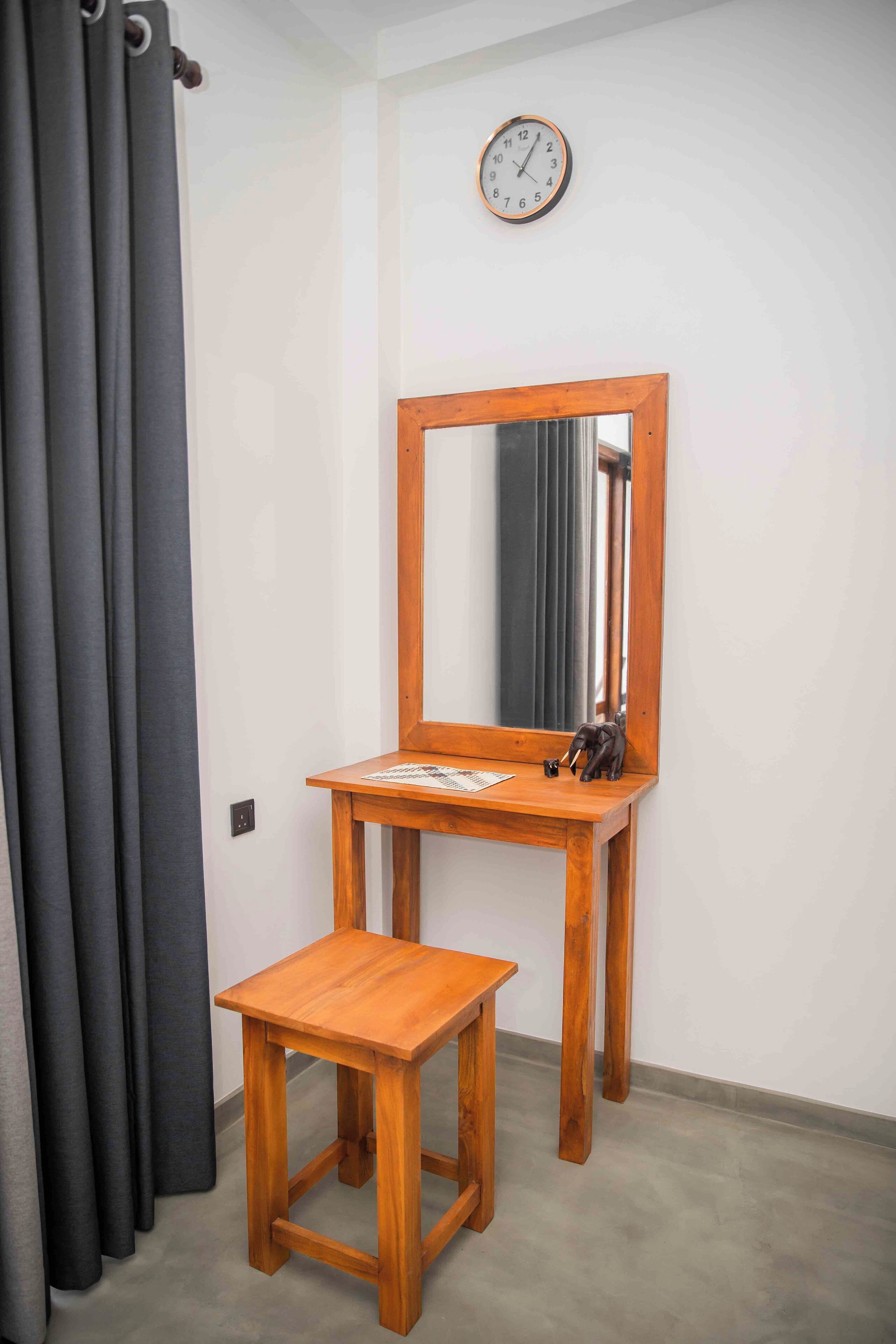 Una habitación con un escritorio y silla de madera, un espejo, un reloj y cortinas grises.