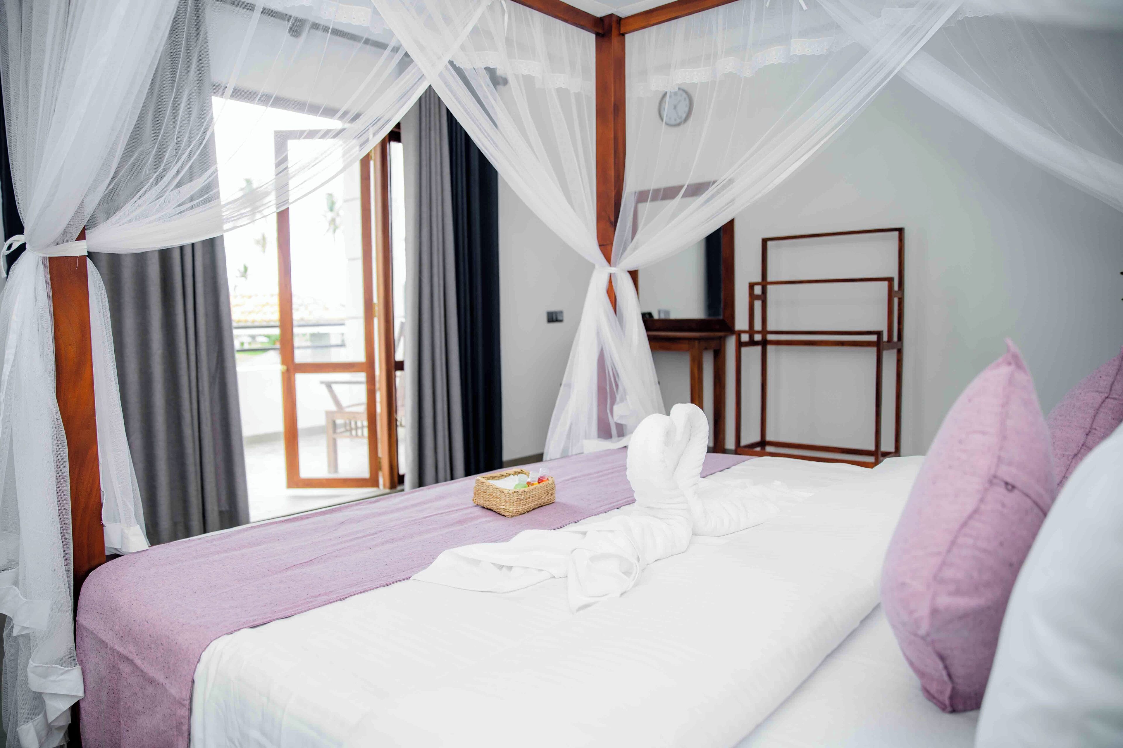 A neatly made bed with a purple bedspread, white pillows, and a white bathrobe. There is a wicker basket on the bed and a wooden chair visible through the window.