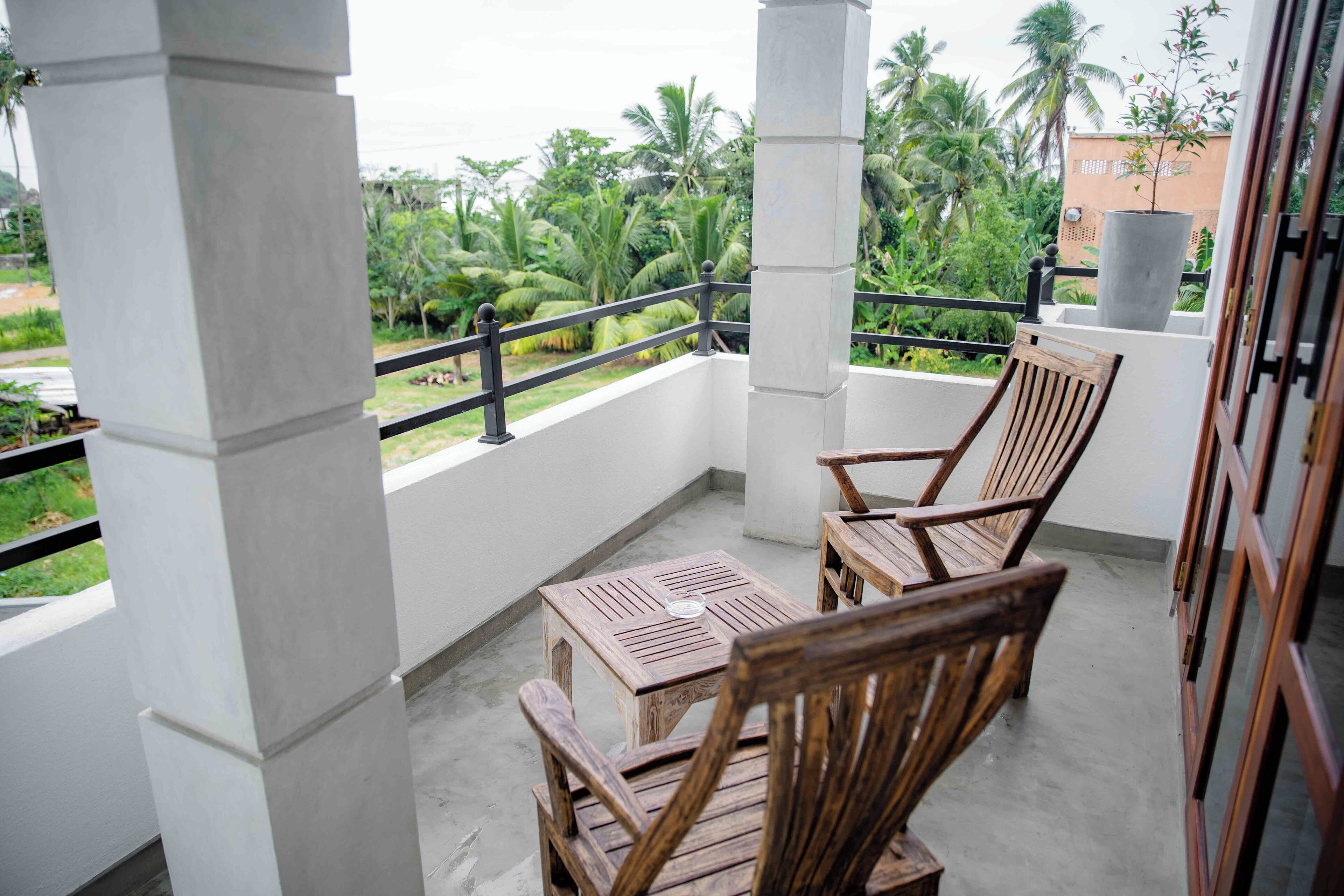 A balcony with wooden chairs and a table, white pillars, and a view of greenery and buildings.