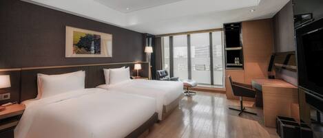 A hotel room with two beds, a desk, a chair, a TV, and a large window with a city view.
