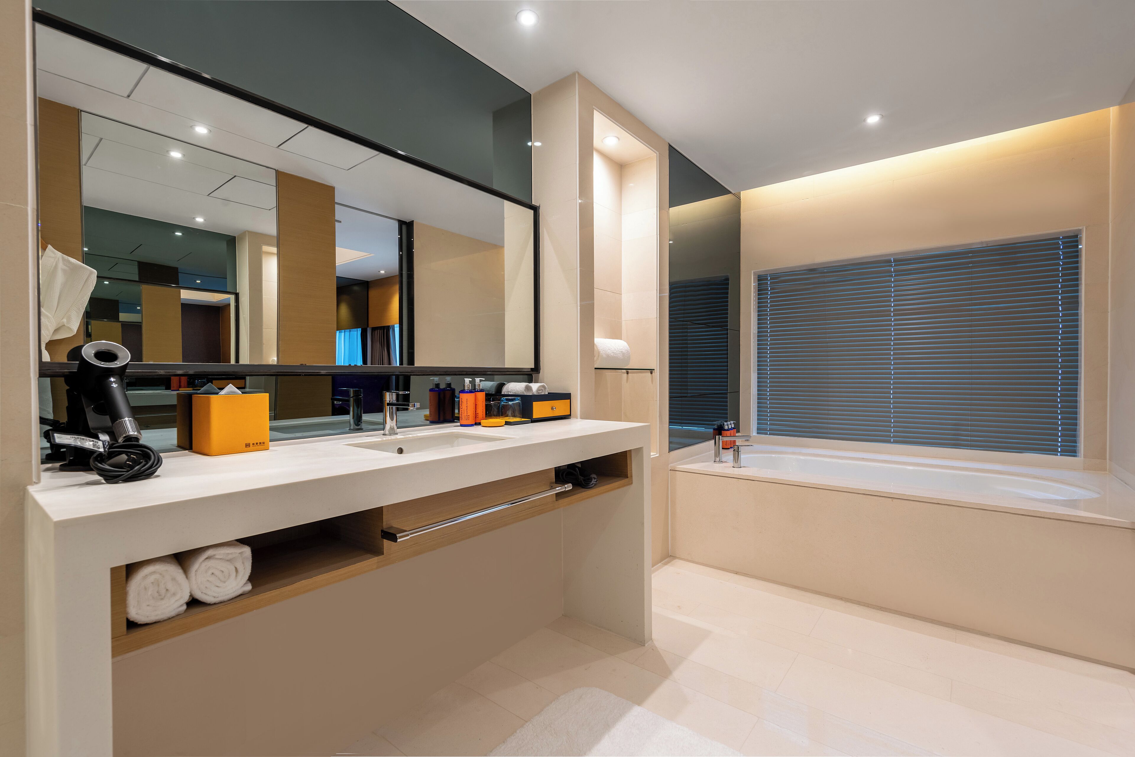 A modern bathroom with a large mirror, dual sinks, a towel rack, and a window with blinds.