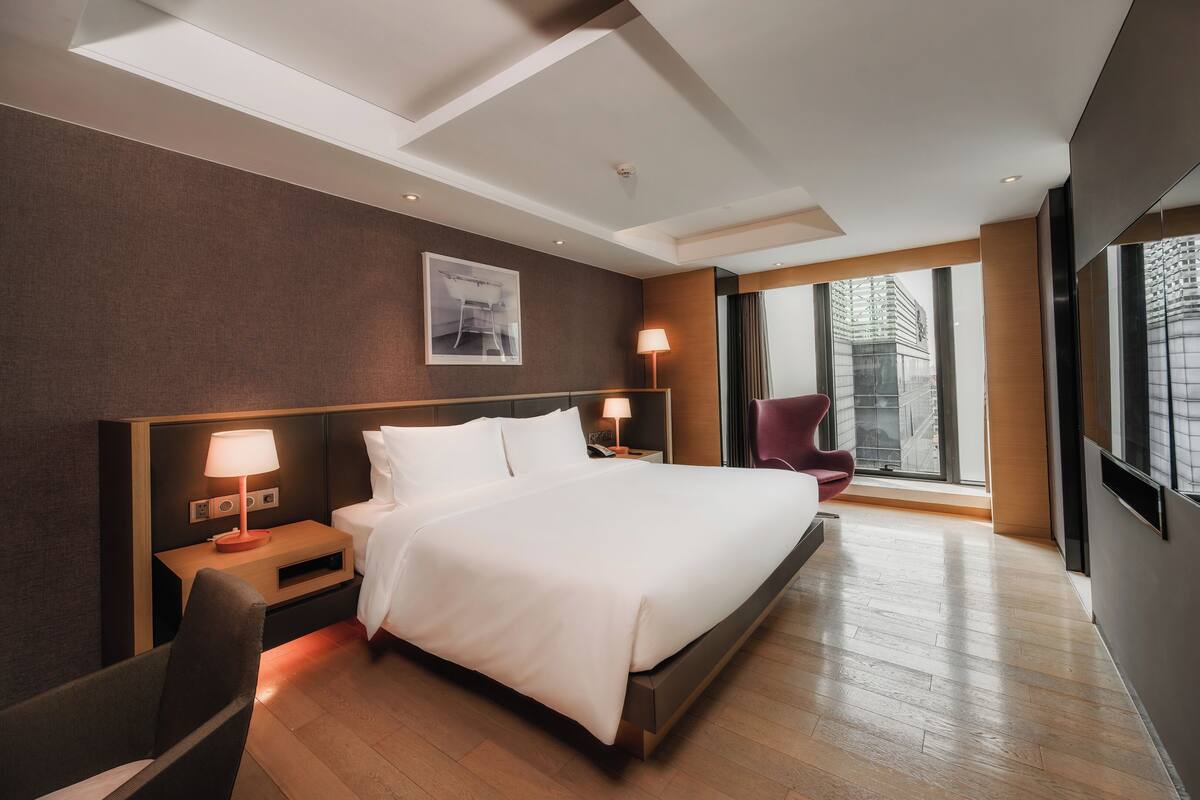 Une chambre d’hôtel moderne dotée d’un grand lit, de tables de chevet avec des lampes, d’un fauteuil et d’une grande fenêtre donnant sur la ville.
