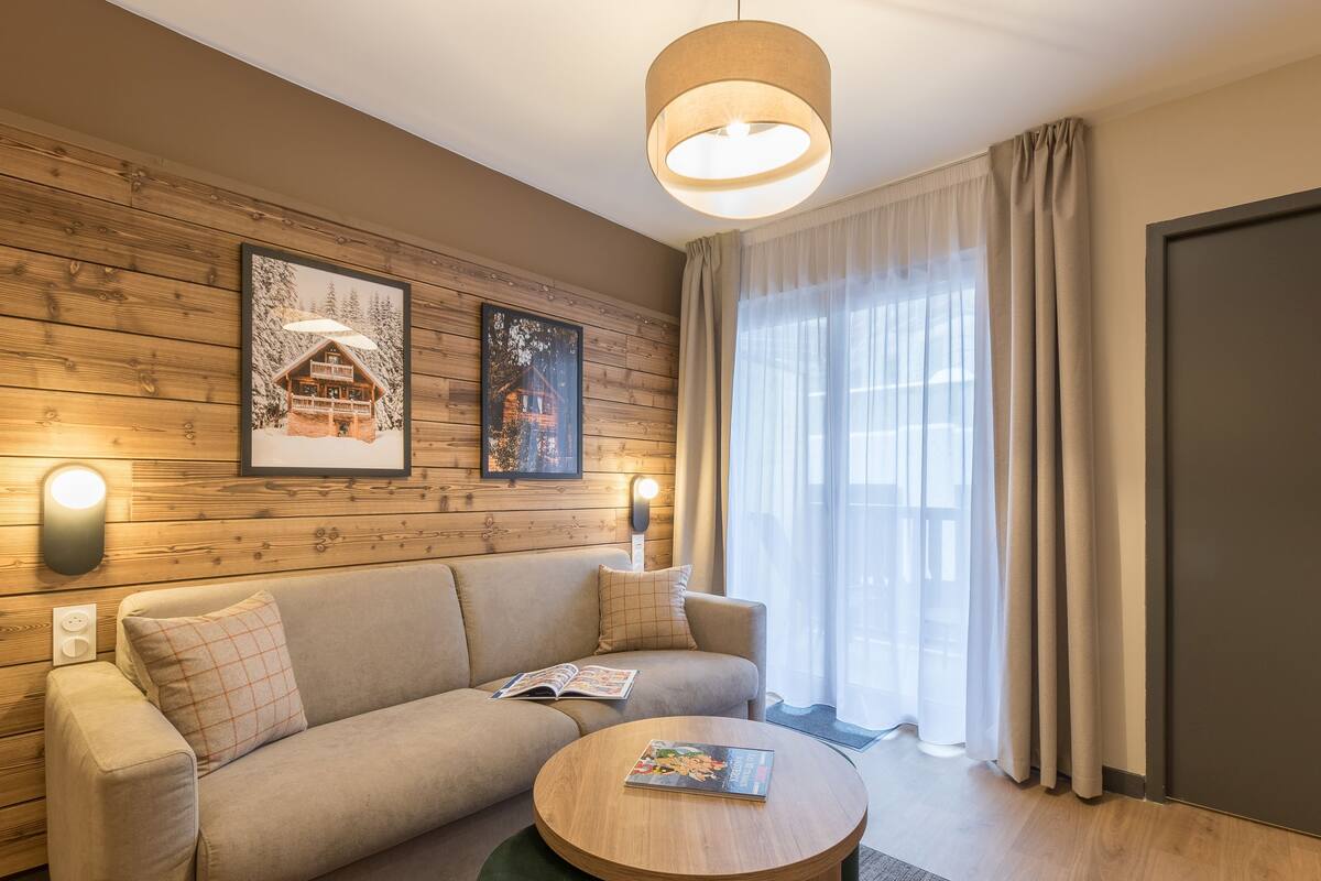 Une pièce chaleureuse avec un mur en bois, un canapé, une table basse ronde et une grande fenêtre avec des rideaux.