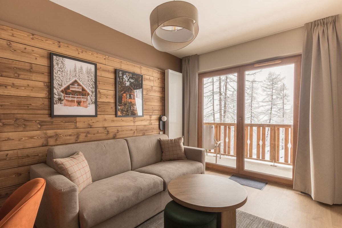 Ein gemütliches Wohnzimmer mit Holzwand, einer Couch, einem runden Tisch und einem Balkon mit Blick auf Bäume.