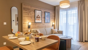 Ein modernes Wohnzimmer mit einer Holzvertäfelung, einer Couch, einem Esstisch mit Frühstücksgebäck und einem großen Fenster mit Vorhängen.