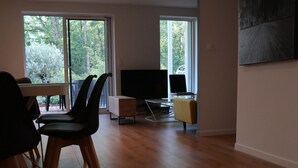 Tilava olohuone, jossa on puulattia, ruokapöytä tuoleineen, televisio ja suuri ikkuna, josta avautuu näkymä puita kohti.