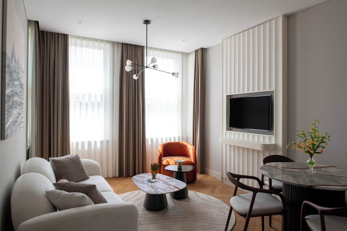 Ein modernes Wohnzimmer mit einer Couch, einem runden Couchtisch, einem Esstisch mit Stühlen, einem Fernseher und einem großen Fenster mit Vorhängen.