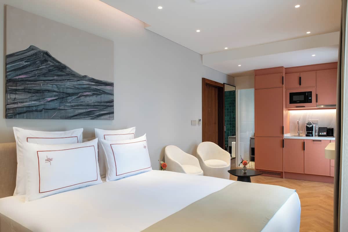 Ein modernes Hotelzimmer mit einem großen Bett, zwei weißen Stühlen, einem kleinen Tisch, einem Bild an der Wand und einer Küchenzeile.