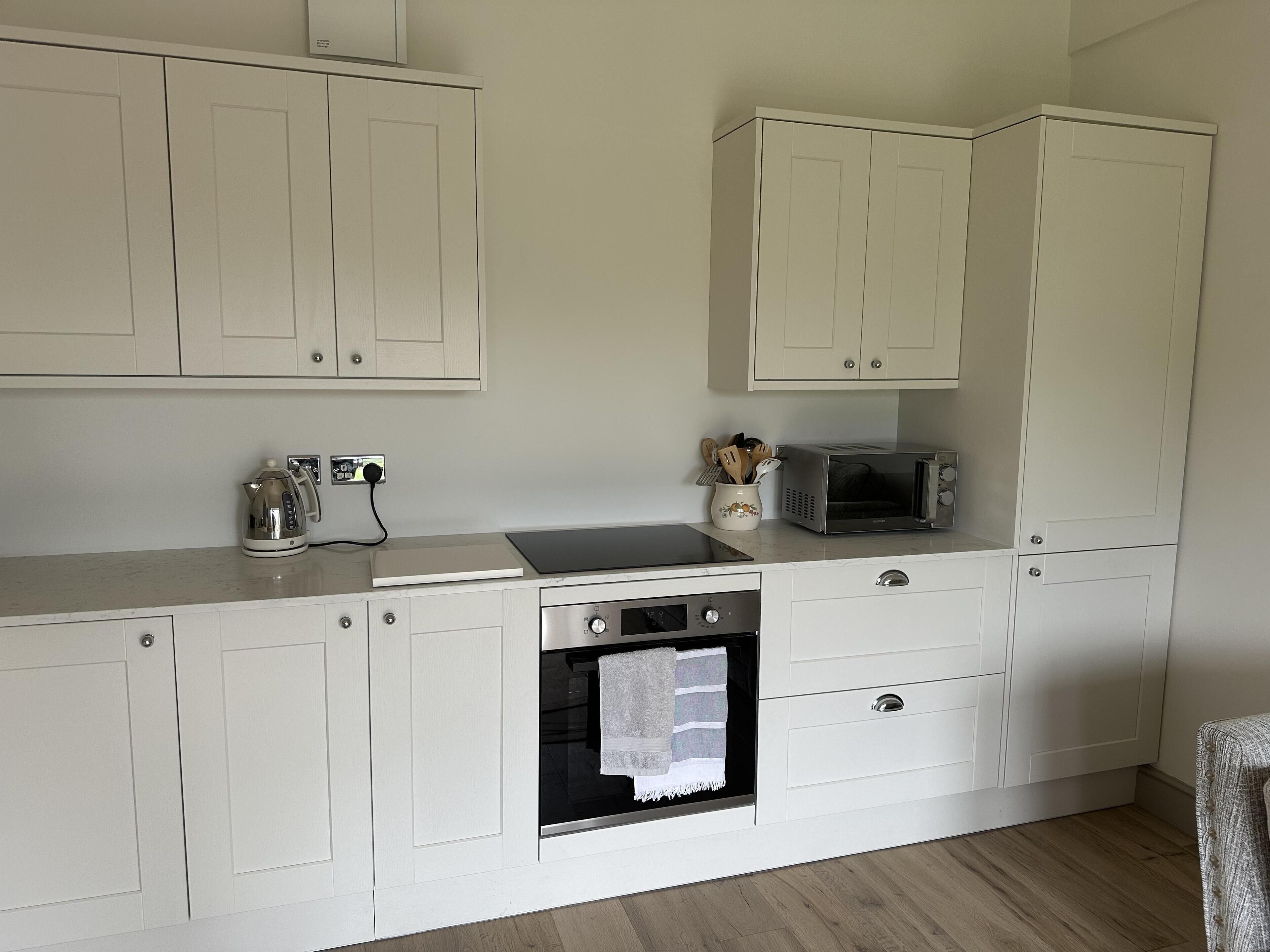 A compact kitchen with white cabinetry, a built-in oven, a microwave, and a kettle on the countertop.