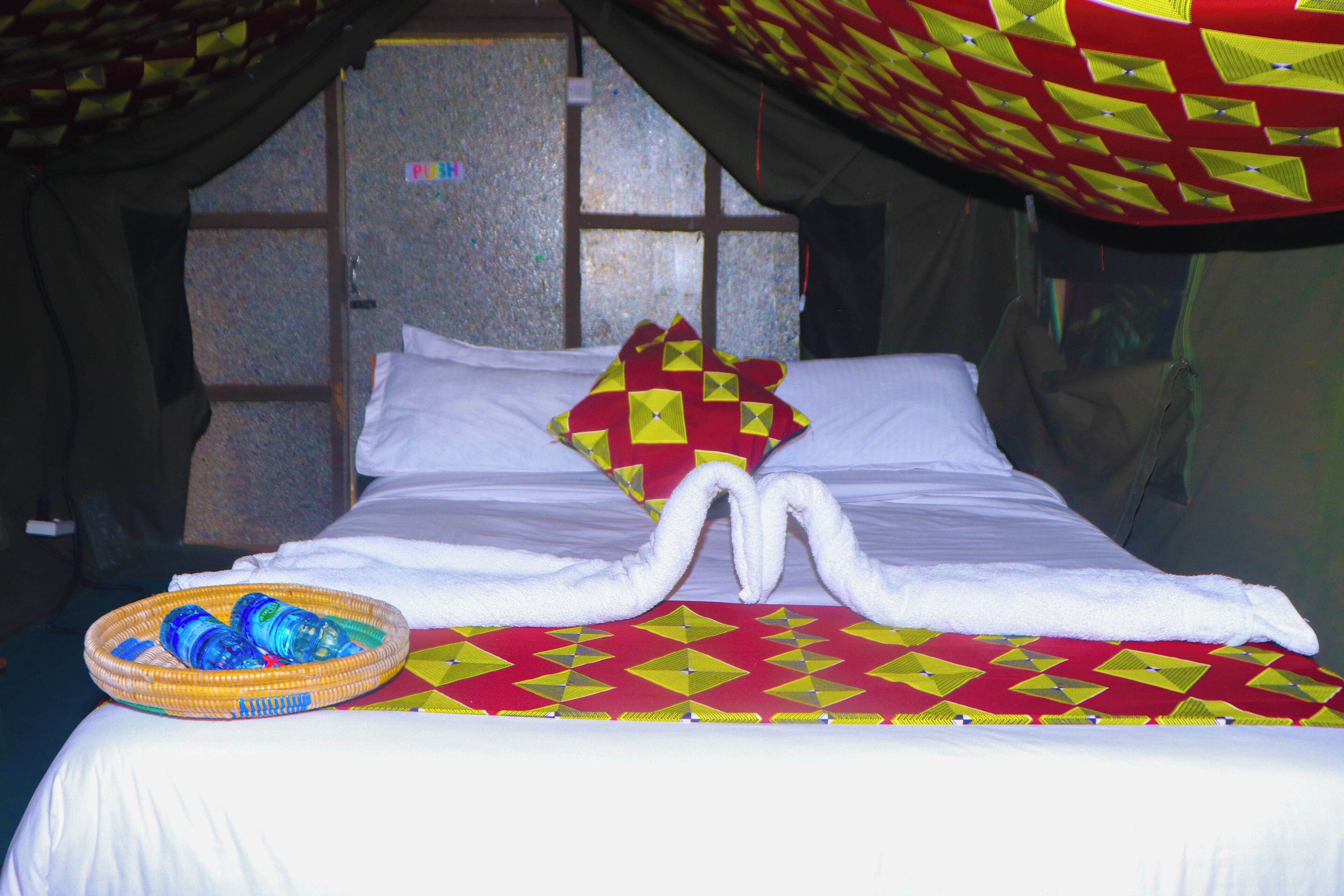 A bed with a red and yellow patterned bedspread, a white pillow, and a towel arranged to resemble two swans. A basket of bottled water is placed on the bed.