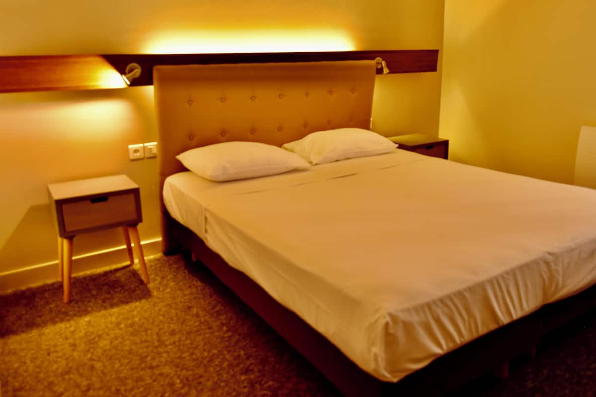 A neatly made bed with a wooden headboard and two pillows, a small bedside table, and a wall-mounted lamp.