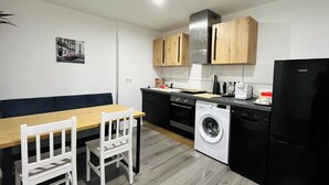 Une cuisine compacte équipée d’un lave-linge, d’un réfrigérateur noir, de meubles en bois et d’un coin repas avec une table et des chaises.
