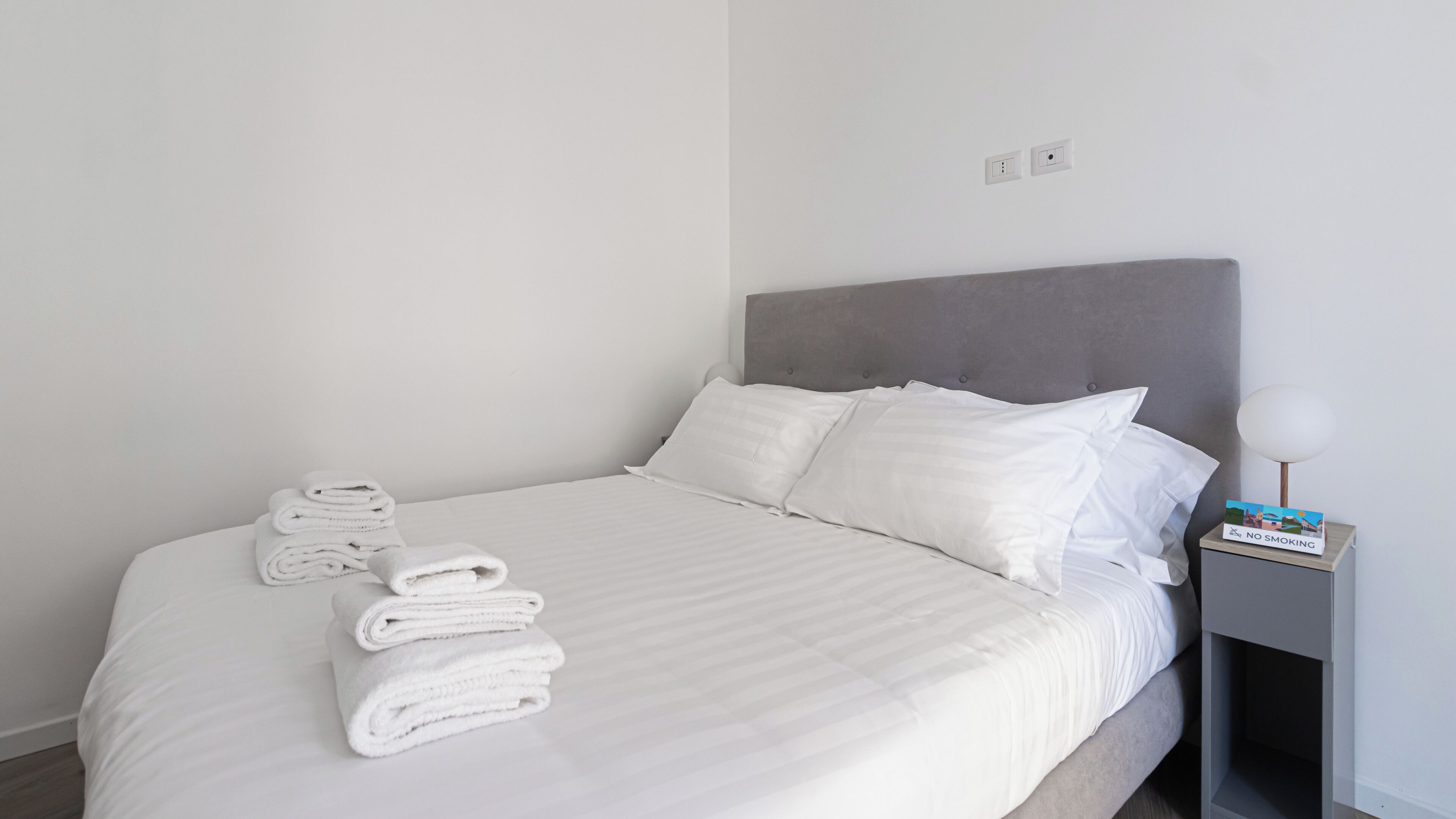 Un lit bien fait, avec du linge de lit blanc et des oreillers, une tête de lit grise, une table de chevet avec une lampe et un panneau indiquant « Il est interdit de fumer ».
