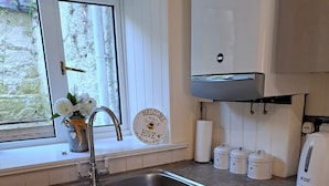 Een keuken met een spoelbak, een raam met uitzicht op groen, een welkomstbord en een waterkoker.