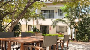An outdoor seating area with a wooden table and chairs, surrounded by greenery and a building with a balcony. - Room in Lodge - Luxury Suite in Boutique Lodge With Free Savannah Adventures 4x4 City Safari (Victoria Falls)