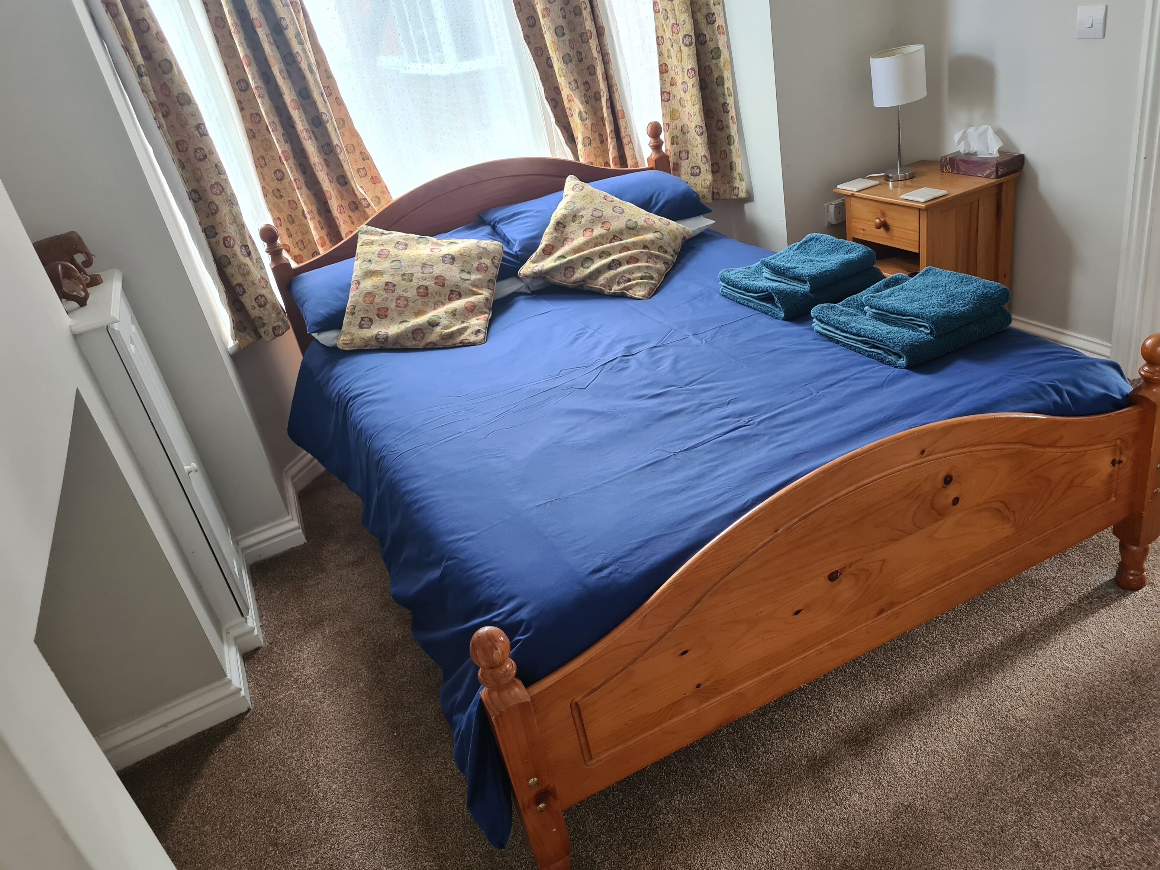 Een slaapkamer met een houten bed, blauw beddengoed, gekleurde kussens, een nachtkastje met een lamp en een zakdoekendoos.