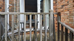 A wooden balcony with a metal railing, a brick wall, and a sliding glass door leading to an interior space with a table and chairs. - Cozy Nottingham Apartment With Perfect Location (Nottingham)
