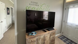 A flat-screen TV on a wooden cabinet with a remote control and a small decorative item. A sign on the wall reads "COME GATHER seashells."
