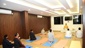 A yoga class in a room with wooden walls, a ceiling with recessed lighting, and a large window. - Arogyadham Wellness Retreat (Rishikesh)