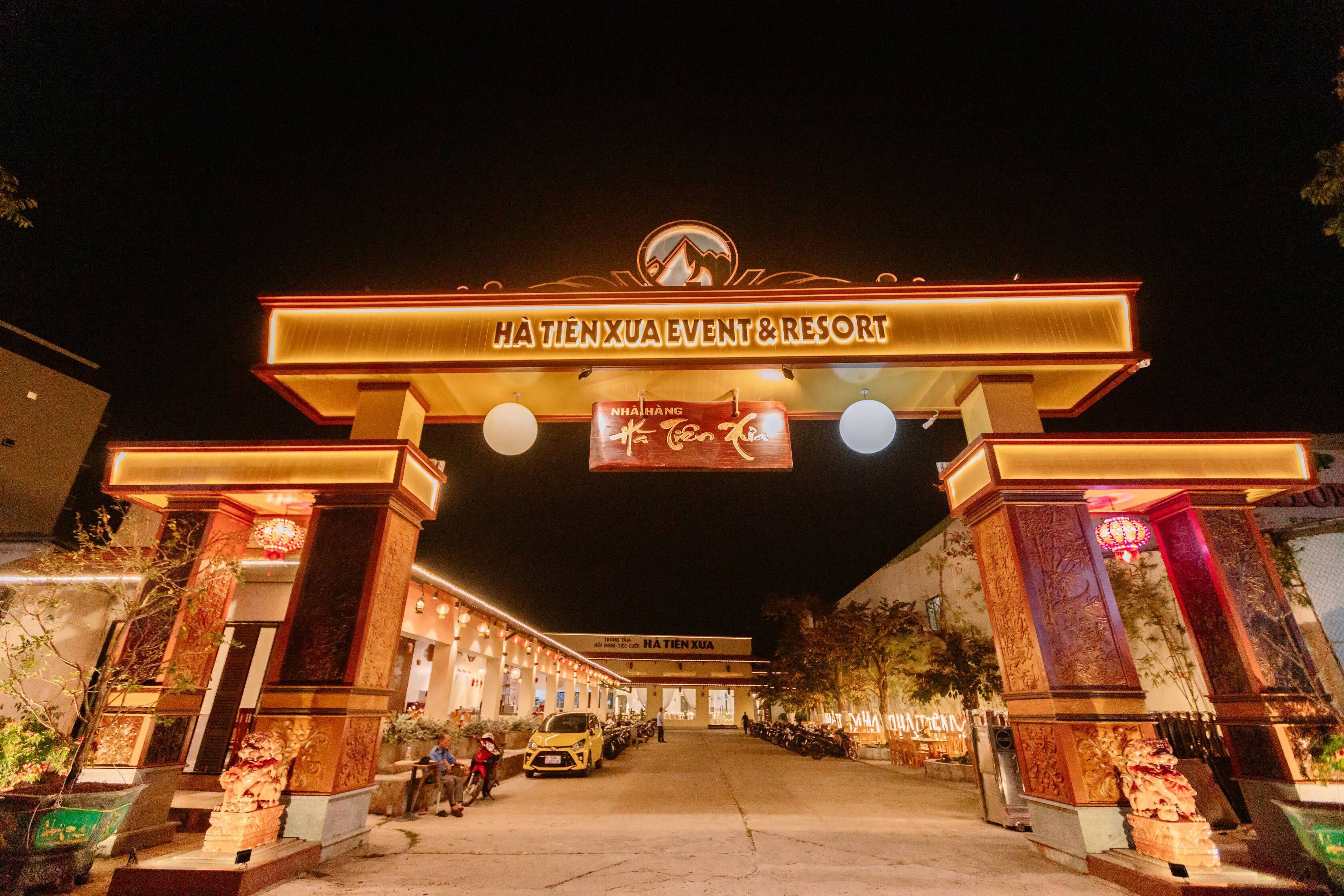 La entrada al Event & Resort Hà Tiên Xua, con un letrero llamativo, una zona de estacionamiento y una fachada decorativa.