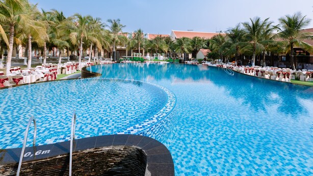 Ein großer, klarer, blauer Pool mit einer gefliesten Kante, umgeben von Palmen und Liegestühlen.