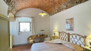 A bedroom with a brick ceiling, a bed with a wooden headboard, a desk with a chair, and a window with curtains.