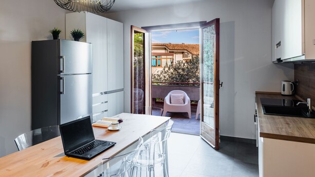 A modern kitchen with a dining area, a laptop on the table, and a view of the outside balcony.
