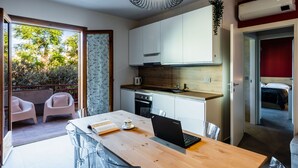 A modern kitchen with white cabinets, a wooden countertop, and a built-in oven. There is a dining area with a table, chairs, and a laptop. A balcony with chairs and a view of trees is visible.