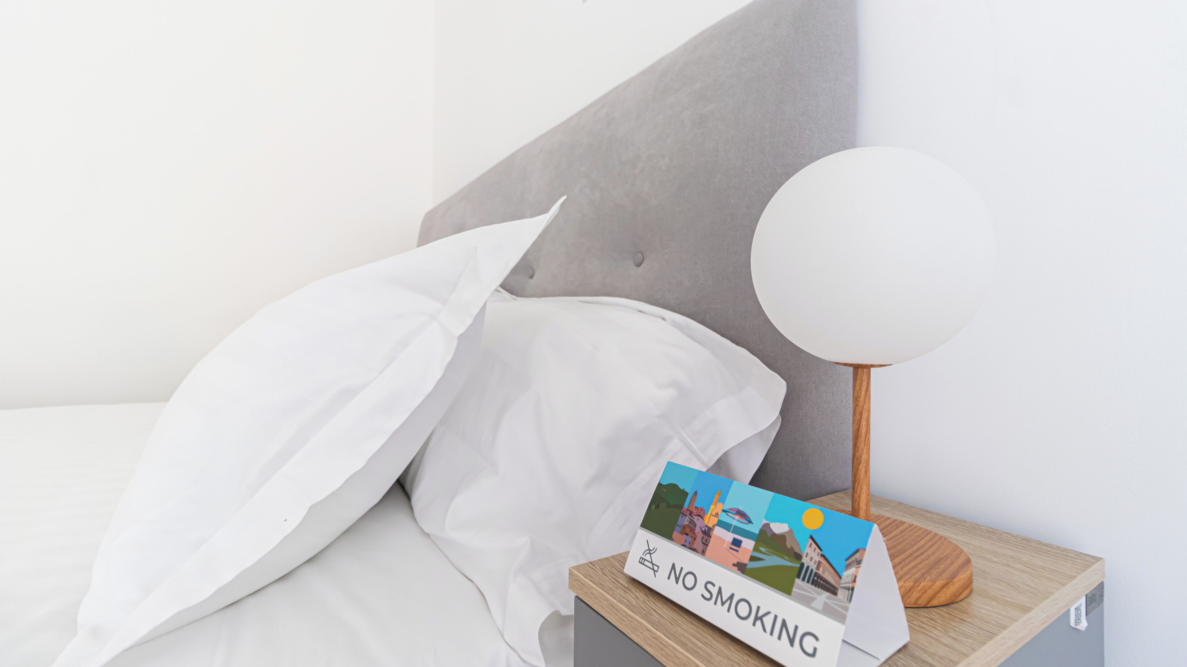 Uma cama bem arrumada com lençóis brancos, cabeceira cinza e mesa de cabeceira com abajur branco e um aviso de proibido fumar.