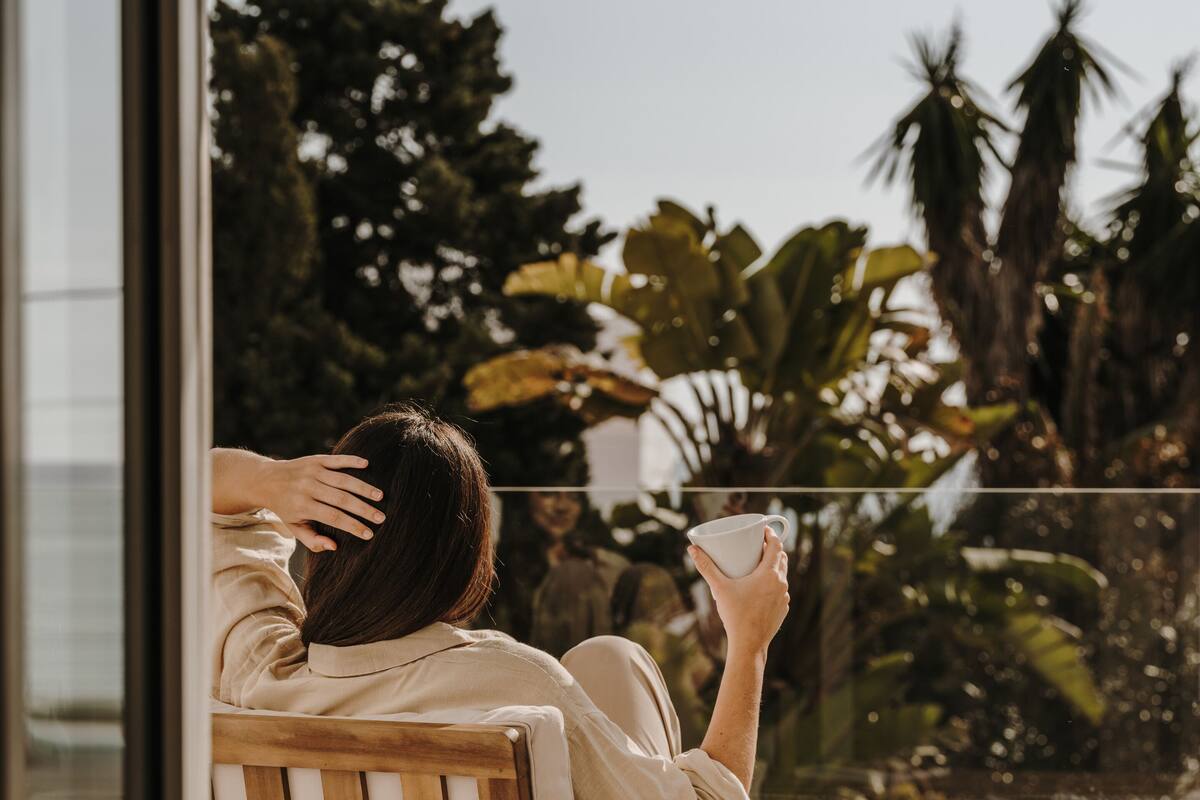 Eine Person sitzt auf einem Holzstuhl mit Kissen, hält eine Tasse und blickt auf einen Balkon mit Blick auf Palmen und ein Gebäude im Hintergrund.