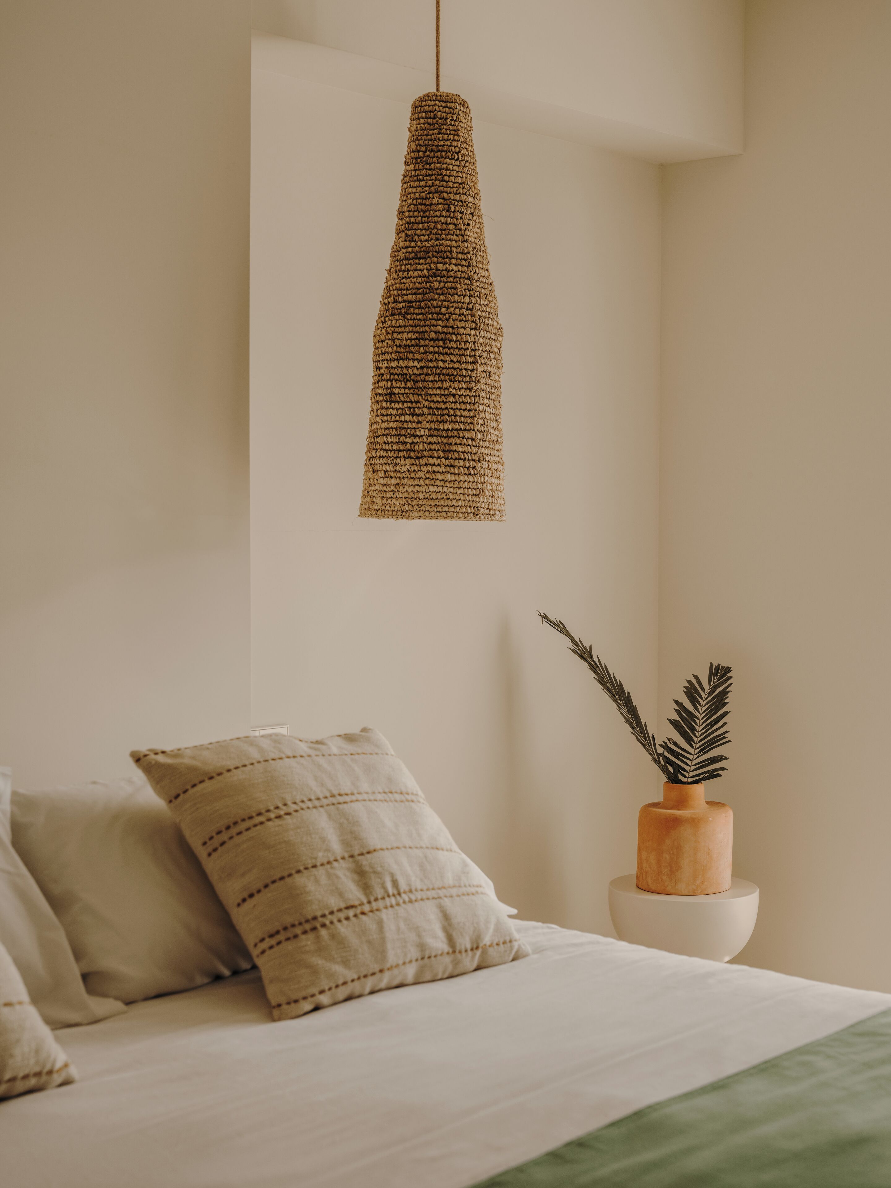 Een slaapkamer met een hangende geweven lamp, een kamerplant in een houten pot en een bed met gestreepte kussens.