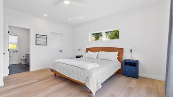 Een slaapkamer met een houten bed, witte wanden, een plafondventilator en een raam met uitzicht op bomen.