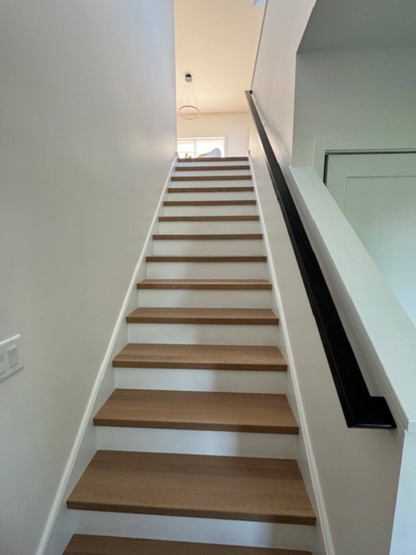 Una escalera con escalones de madera y paredes blancas que conduce a un rellano con barandilla de vidrio.