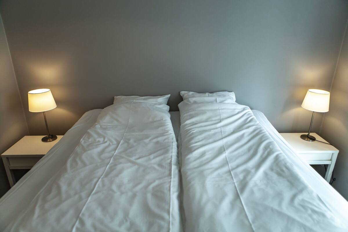 Un lit simple avec une literie blanche, dans une pièce aux murs gris. Il y a deux tables de chevet, chacune avec une lampe.