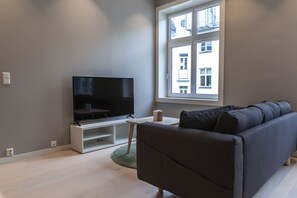Een moderne woonkamer met een flatscreen televisie op een witte standaard, een donkergrijze bank en een kleine witte tafel met een roze kaars.