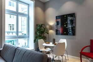 Una sala de estar moderna con un sofá gris, un área de comedor con sillas blancas y una mesa, una planta en maceta y un cuadro en la pared.
